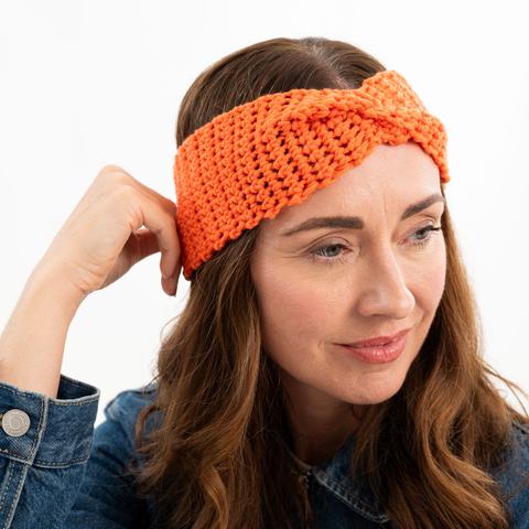 Image of a smiling person in quarter profile resting their hand against their head. They're wearing a denim jacket and a bright orange crochet headband with a twist detail at the front.