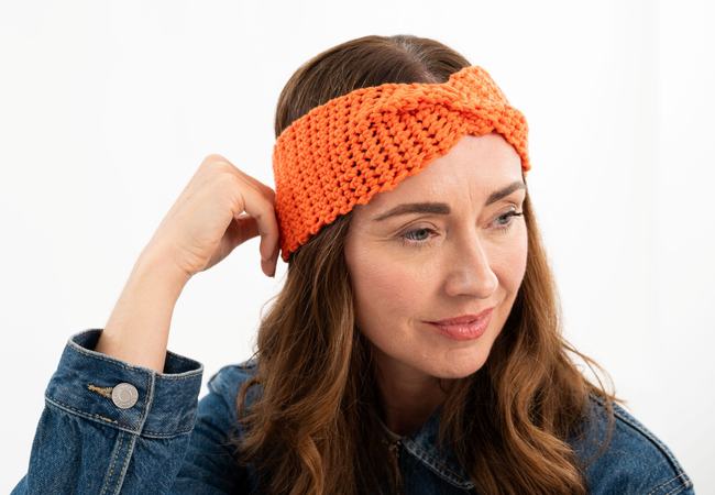Image of a smiling person in quarter profile resting their hand against their head. They're wearing a denim jacket and a bright orange crochet headband with a twist detail at the front.