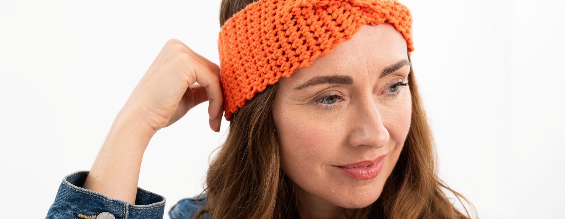 Image of a smiling person in quarter profile resting their hand against their head. They're wearing a denim jacket and a bright orange crochet headband with a twist detail at the front.
