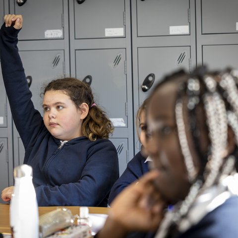 A schoolgirl raises her hand in a UK primary school classroom.