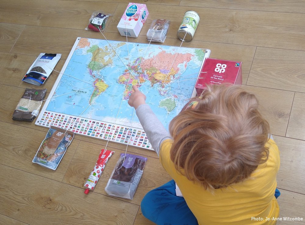 A child looks at a map of the world with pins that strings to bags of real food from their family cupboard