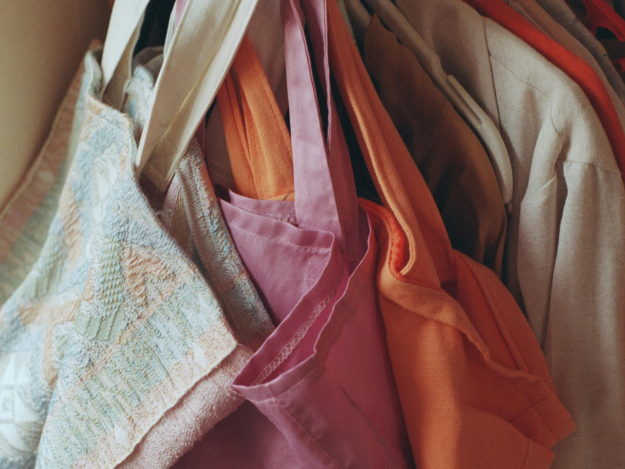 3 colourful tote bags in the colours orange, pink and one multi-colour pastels. They are hanging on a clothing rack placed in front of some clothing.