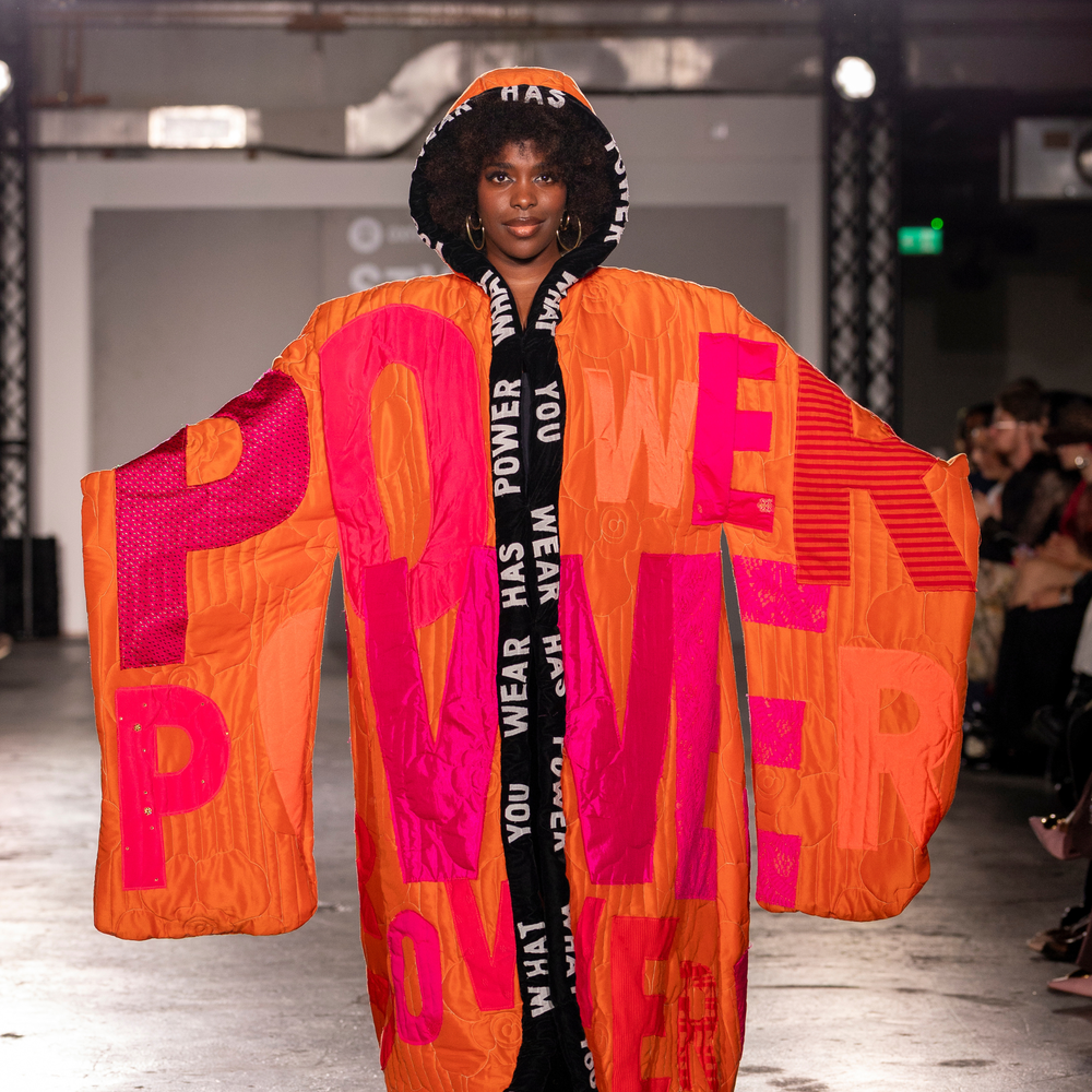 Sabrina Grant wearing a colourful cape on the catwalk at the Oxfam x Vinted Style for Change fashion show. The cape is emblazoned with the slogan: 'What you wear has power'.