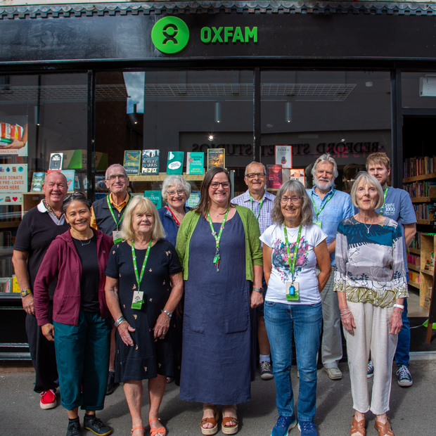 Group photo of Oxfam volunteers and staff standing outside an Oxfam bookshop, beneath the green Oxfam storefront sign. Shop window displays promote donating and volunteering, while a signboard advertises the store’s grand opening.