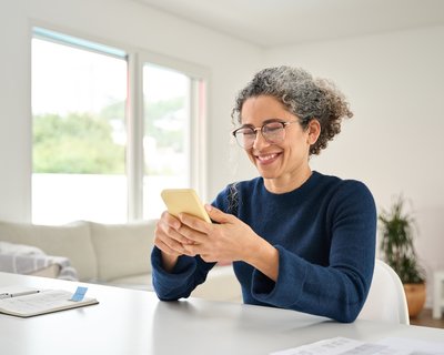 A woman with curly grey and brown hair, wearing glasses and a navy blue sweater, smiles while looking at her smartphone.