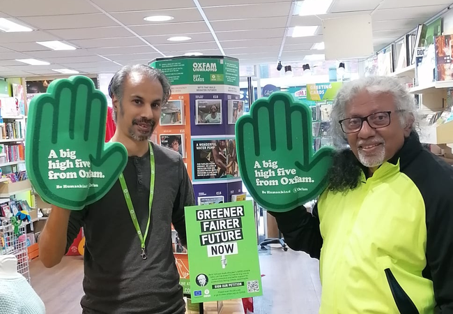 Vimal and Push in an Oxfam shop wearing green foam hands that say 'a big high five from Oxfam' with a 'greener, fairer future now' poster in the background.
