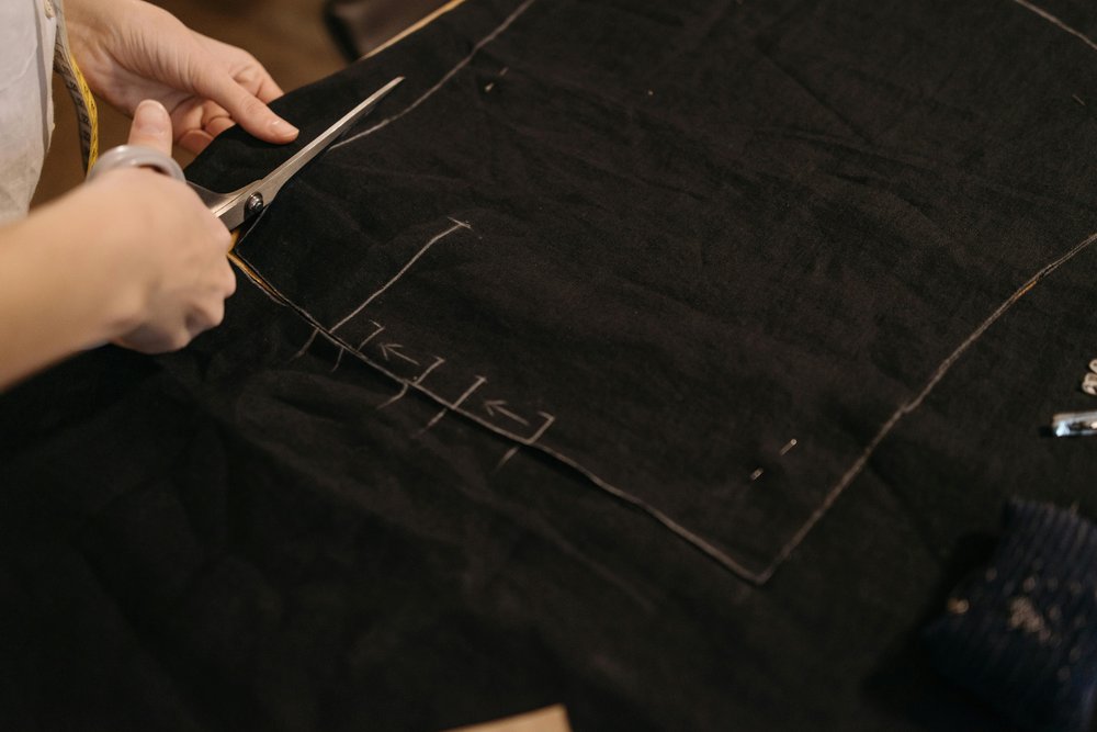 A black piece of clothing/ fabric with a pair of hands with scissors cutting the material.