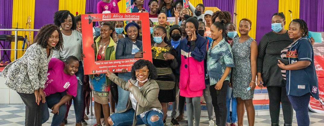 Young Kenyan women in a group photo with some holding large cardboard picture frame cutouts and peering through the gap.