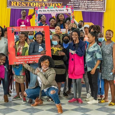 Young Kenyan women in a group photo with some holding large cardboard picture frame cutouts and peering through the gap.