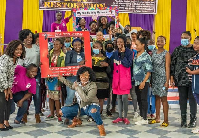 Young Kenyan women in a group photo with some holding large cardboard picture frame cutouts and peering through the gap.