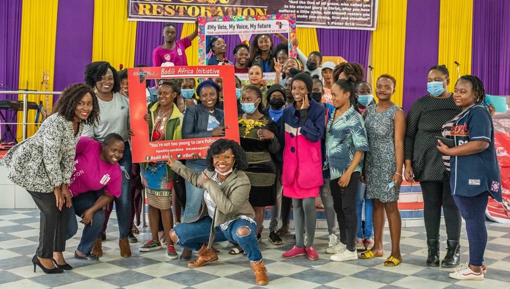 Young Kenyan women in a group photo with some holding large cardboard picture frame cutouts and peering through the gap.