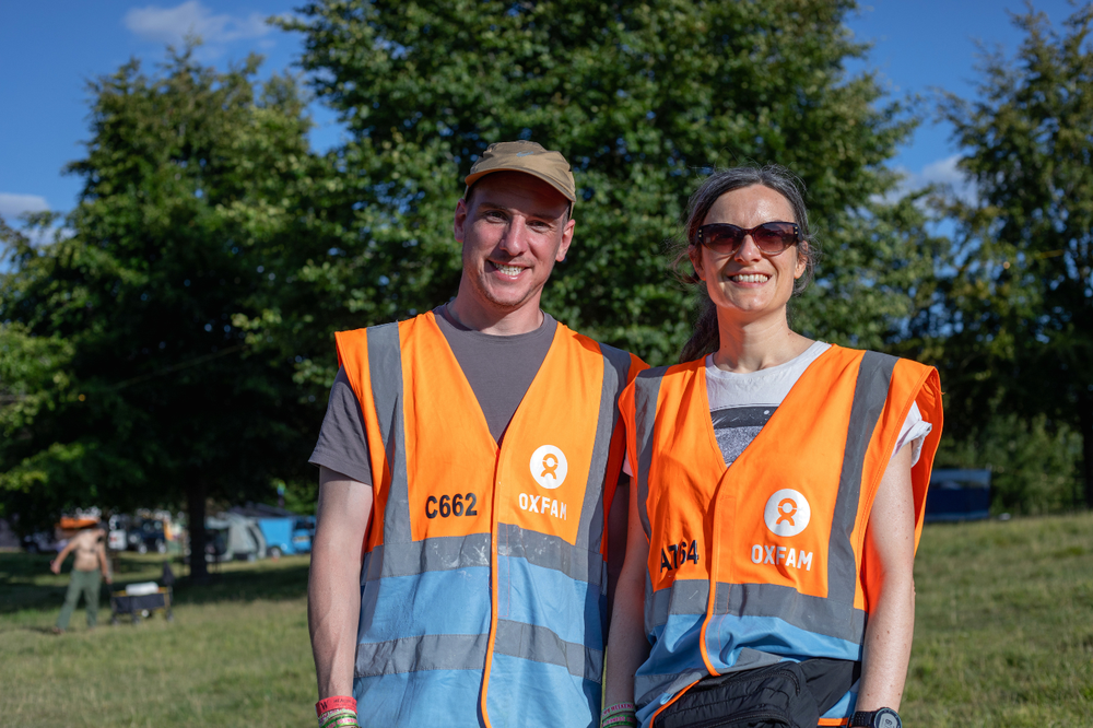 Two Oxfam festival volunteers smiling at the camera with a line of trees behind them