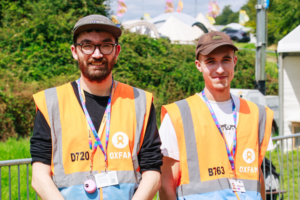 Two Oxfam volunteer stewards smiling at the camera with festival stages and flags in the background