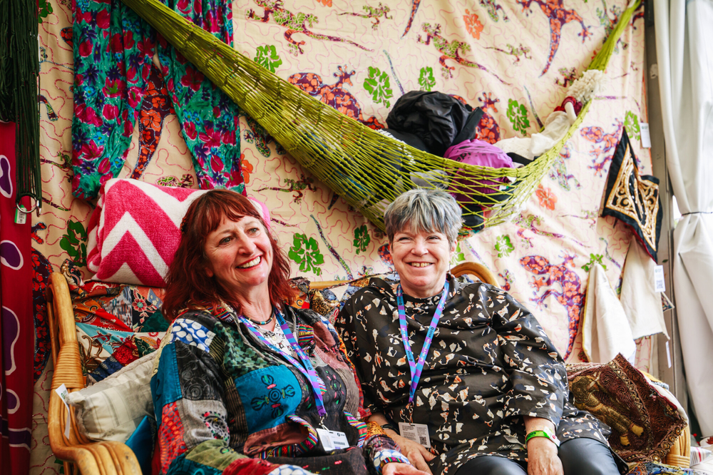 Two Oxfam festival shop volunteers sitting in a shop on a sofa with draped patterned fabrics in the background