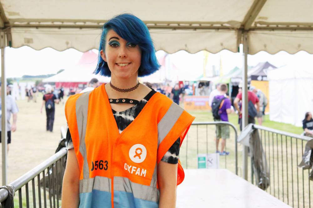 Oxfam festival steward with blue hair smiling at the camera while working on a festival gate