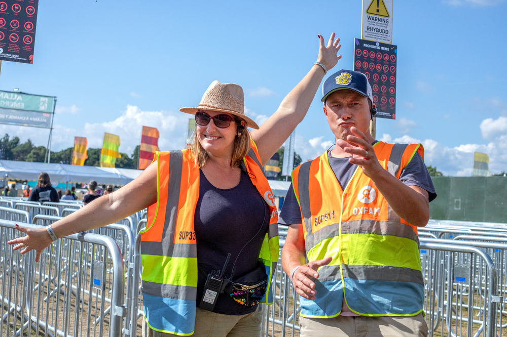 Two Oxfam stewards standing at a festival gate, one with arms in the air and the second playing an air trumpet