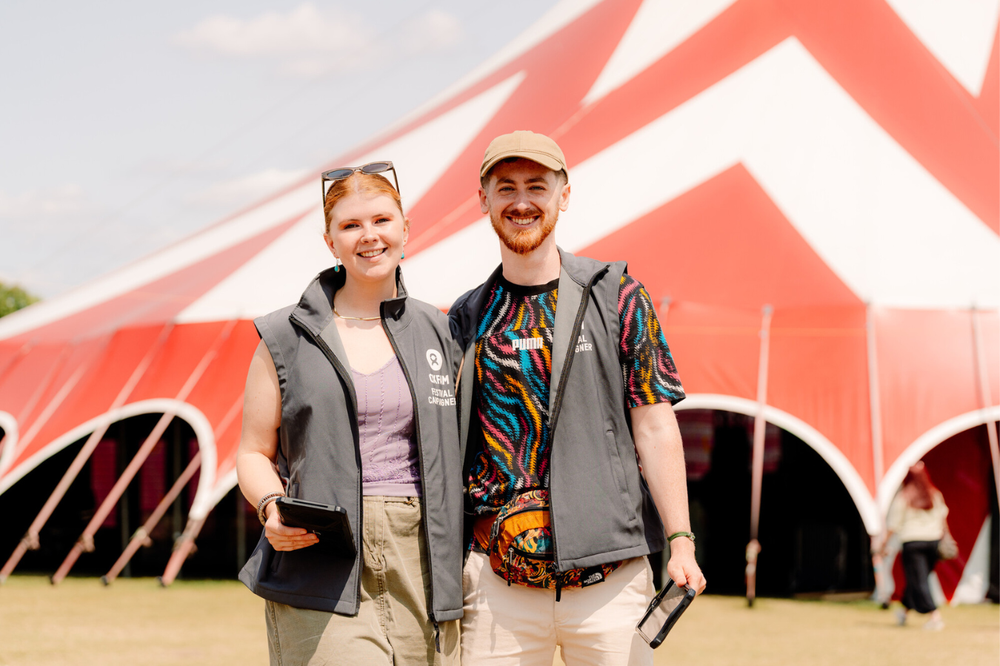Two Oxfam Festival Campaigners holding ipads and smiling at the camera. Behind them is a red and white stripey festival stage tent