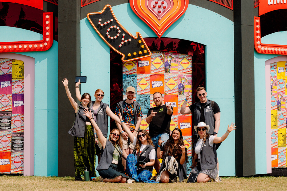 Group of Oxfam festival campaigners in front of a colourful Mighty Hoopla installation including a giant heart and arrow with the words 'joy' written on it