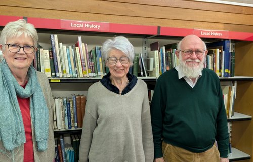 Three people standing in front of bookshelves labelled “Local History” in a library setting, with rows of books visible behind them.