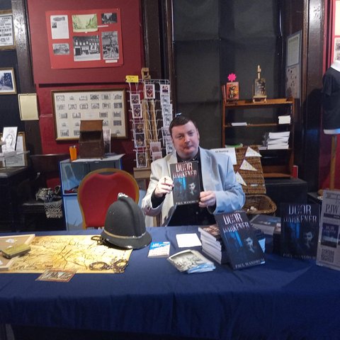 Paul sitting behind a book signing table, holding a copy of his book