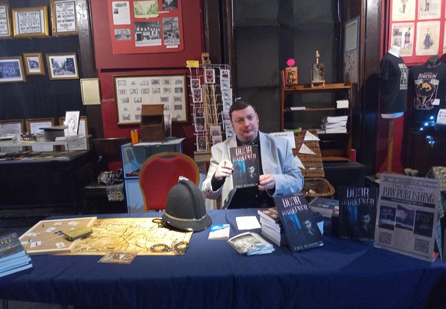 Paul sitting behind a book signing table, holding a copy of his book