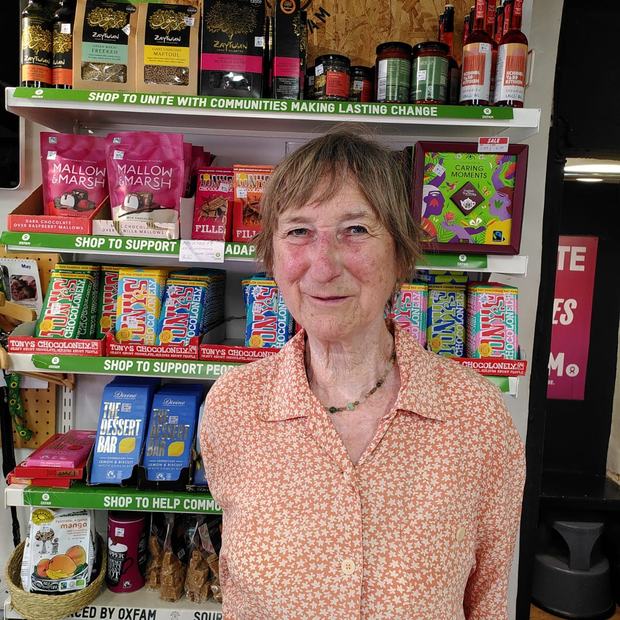 A person wearing a patterned orange shirt stands in front of a “Sourced by Oxfam” display stocked with chocolate bars, snacks, preserves and beverages.