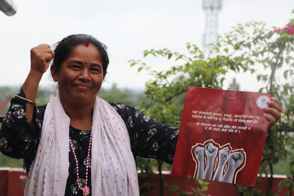 A smiling woman raises her fist in solidarity while holding a red placard with text in Nepali and illustrations of raised fists, standing outdoors near green foliage.