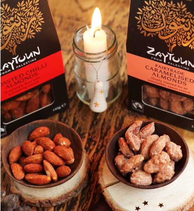 Two small wooden bowls, both holding two different flavours of almonds with the package behind, the packet is brown, long and has a gold tree. In the Centre of the packages is a lit candle sitting in a clear jar.