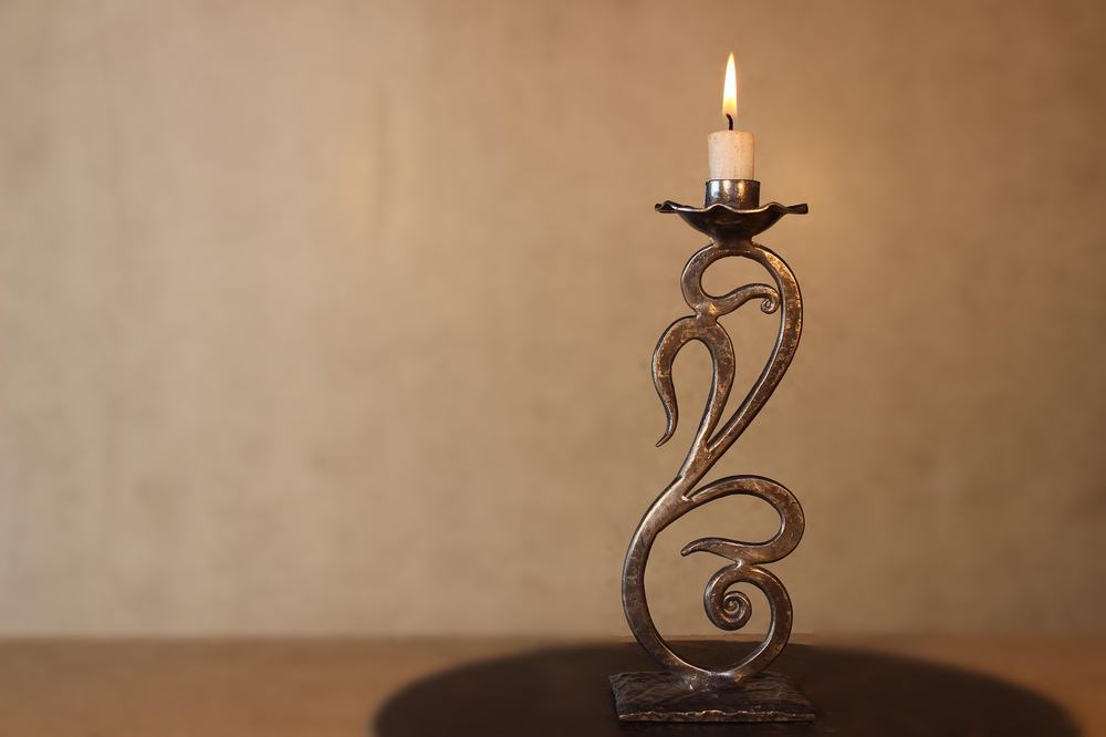 A bronze candlestick holder with a swirly design sits on a dark wood table. In the candlestick is a lit candle.
