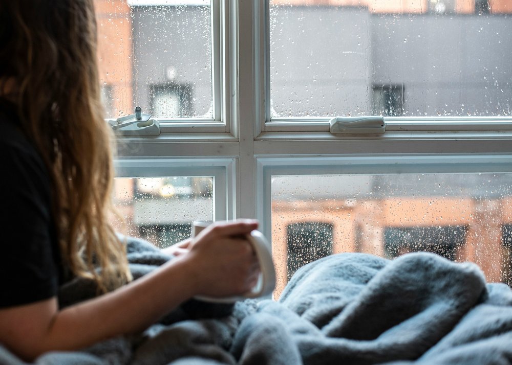 Woman with long hair holding a mug, under a blanket and looking out of a window with rain on it