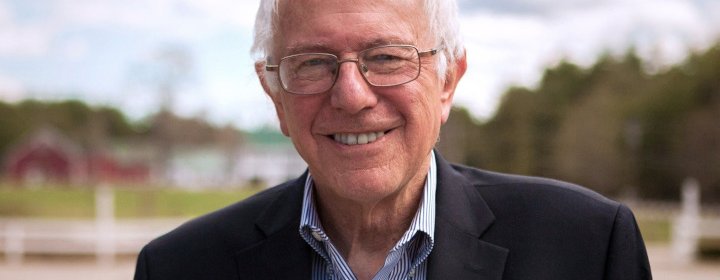 Senator Bernie Sanders wearing a dark suit with a light blue and white striped shirt. He has white hair, wears glasses and is smiling. The background shows some dark red buildings, white buildings with green roofs, trees and there are clouds in the sky.