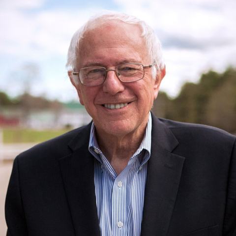 Senator Bernie Sanders wearing a dark suit with a light blue and white striped shirt. He has white hair, wears glasses and is smiling. The background shows some dark red buildings, white buildings with green roofs, trees and there are clouds in the sky.