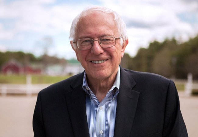 Senator Bernie Sanders wearing a dark suit with a light blue and white striped shirt. He has white hair, wears glasses and is smiling. The background shows some dark red buildings, white buildings with green roofs, trees and there are clouds in the sky.