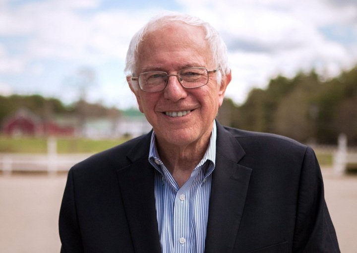 Senator Bernie Sanders wearing a dark suit with a light blue and white striped shirt. He has white hair, wears glasses and is smiling. The background shows some dark red buildings, white buildings with green roofs, trees and there are clouds in the sky.