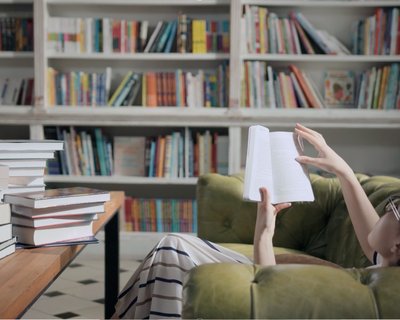Person lying on a green sofa reading a book, with a wooden table stacked with books in the foreground and shelves filled with colourful books in the background.