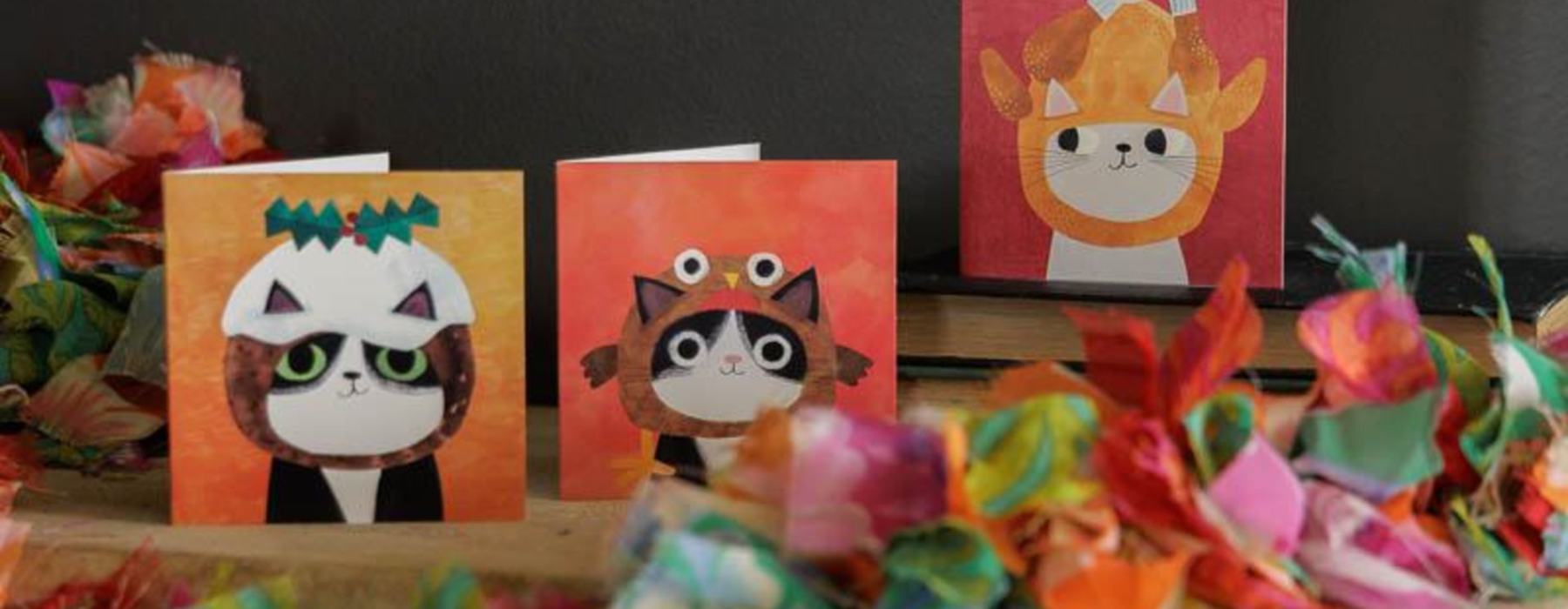 Three fun Xmas cards on a mantelpiece covered in colourful decorations. A cartoon cat is on each one. The first cat wears a Christmas pudding hood, a second cat wears a hood in the shape of a roast turkey and the third cat wears a robin hat.