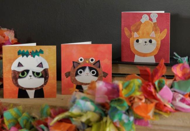 Three fun Xmas cards on a mantelpiece covered in colourful decorations. A cartoon cat is on each one. The first cat wears a Christmas pudding hood, a second cat wears a hood in the shape of a roast turkey and the third cat wears a robin hat.