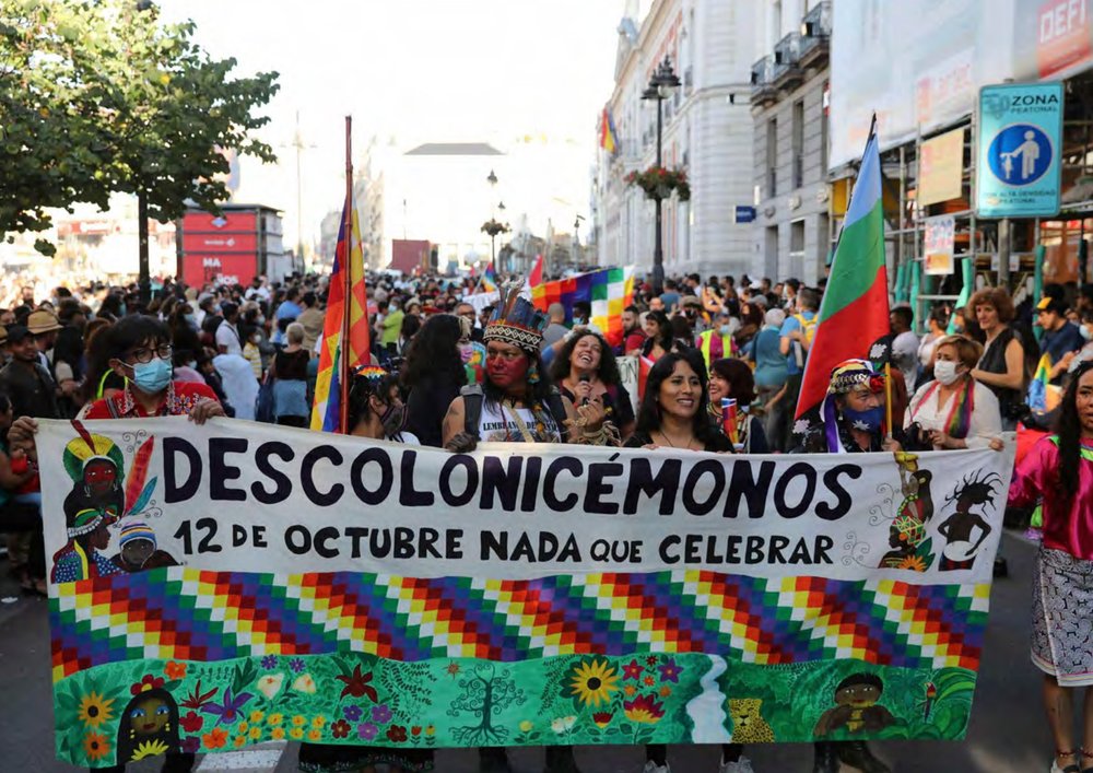 A demonstration on the street of a Latin American city with men and women smiling, waving colourful flags and holding a very large colourful banner with the Spanish words "Descolonicémonos 12 de Octubre nada que celebrar".