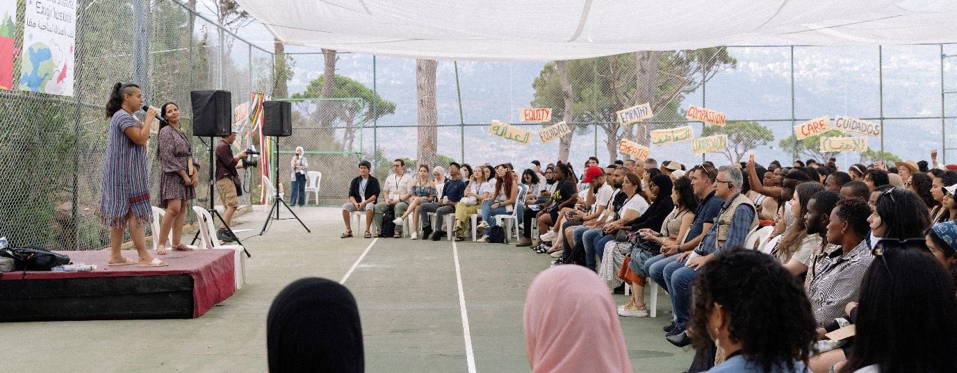 Two people are stood on a small stage speaking to a sitting audience of a diverse group of people. Around the edge of the seating area are banners with single words in English, Spanish and Arabic: equity, care, empathy, compassion.