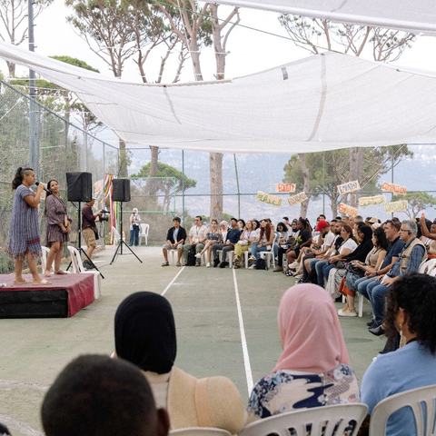Two people are stood on a small stage speaking to a sitting audience of a diverse group of people. Around the edge of the seating area are banners with single words in English, Spanish and Arabic: equity, care, empathy, compassion.