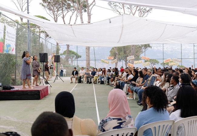 Two people are stood on a small stage speaking to a sitting audience of a diverse group of people. Around the edge of the seating area are banners with single words in English, Spanish and Arabic: equity, care, empathy, compassion.