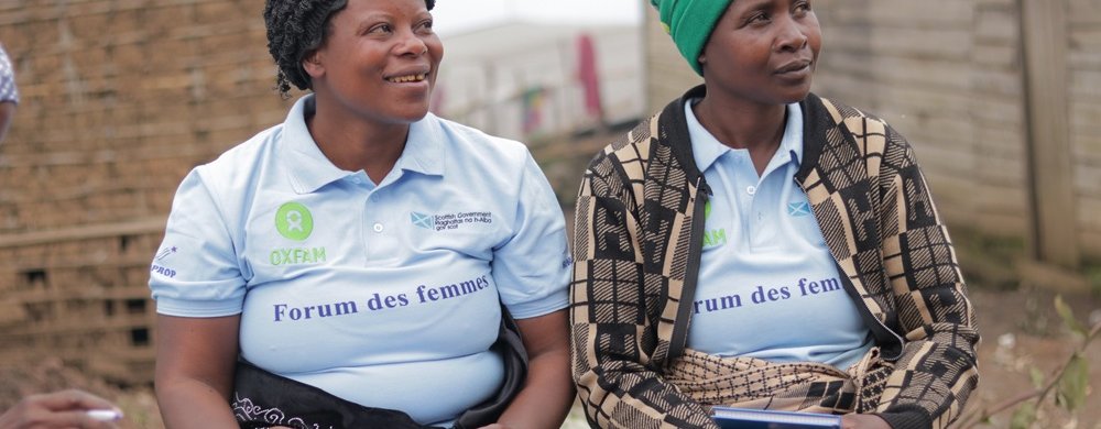 Two women sit side by side outdoors, smiling and listening during a community meeting. Both wear light blue Oxfam-branded “Forum des femmes” polo shirts. One woman has a black headscarf and holds papers, while the other wears a green knitted hat and a pat