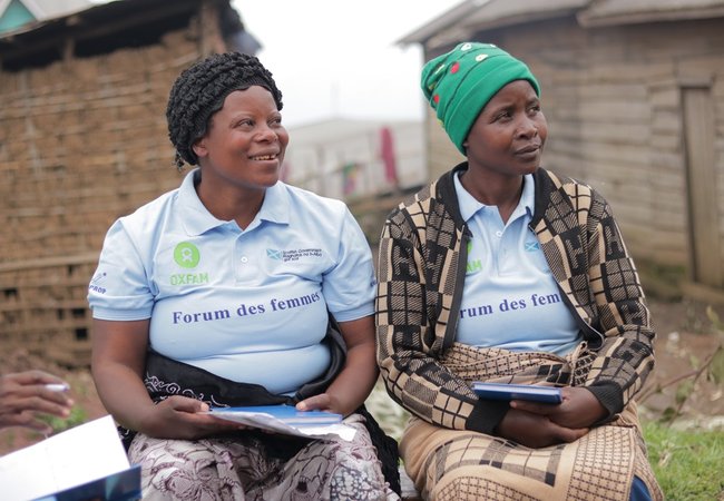 Two women sit side by side outdoors, smiling and listening during a community meeting. Both wear light blue Oxfam-branded “Forum des femmes” polo shirts. One woman has a black headscarf and holds papers, while the other wears a green knitted hat and a pat