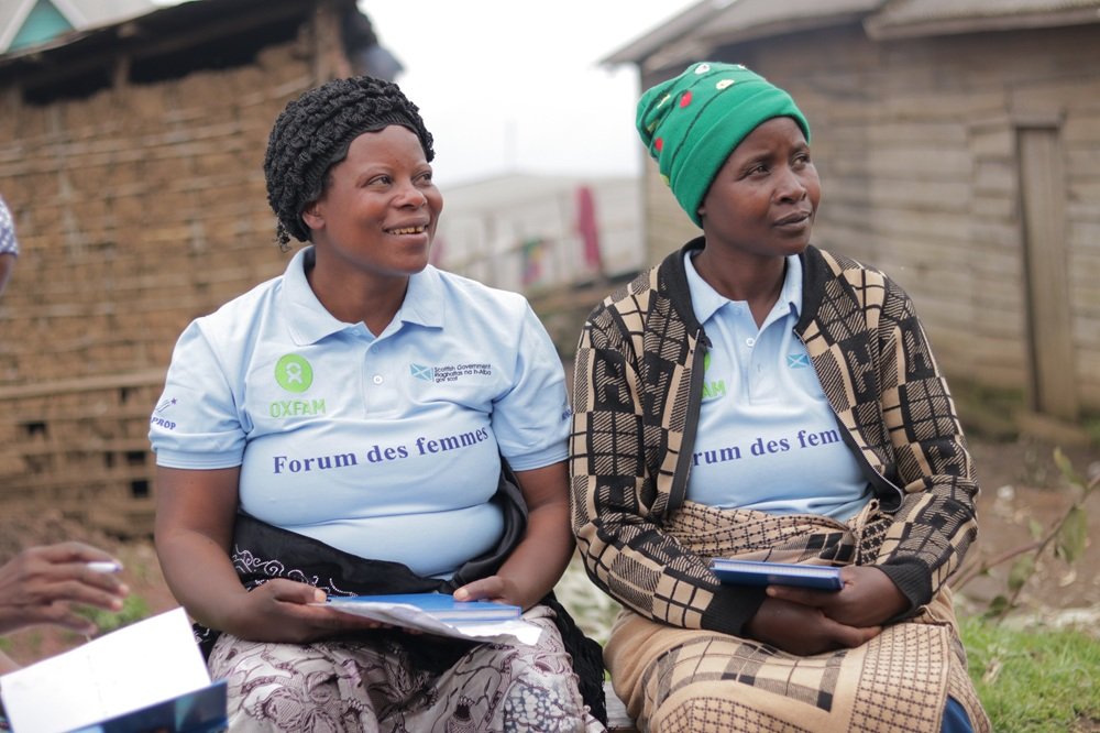 Two women sit side by side outdoors, smiling and listening during a community meeting. Both wear light blue Oxfam-branded “Forum des femmes” polo shirts. One woman has a black headscarf and holds papers, while the other wears a green knitted hat and a pat