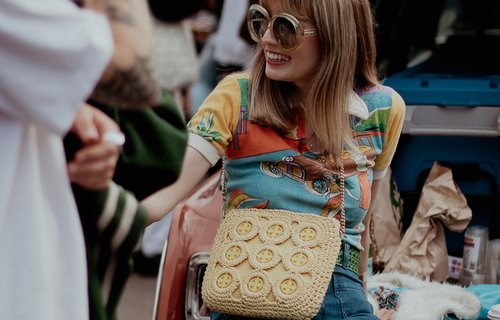 A woman sat on the back of a red truck wearing a vintage outfit. She wears a multi-coloured red, yellow, and blue t-shirt with a collar,  fairly large pair of circle sunglasses with blue jeans and a cream strap-over bag.