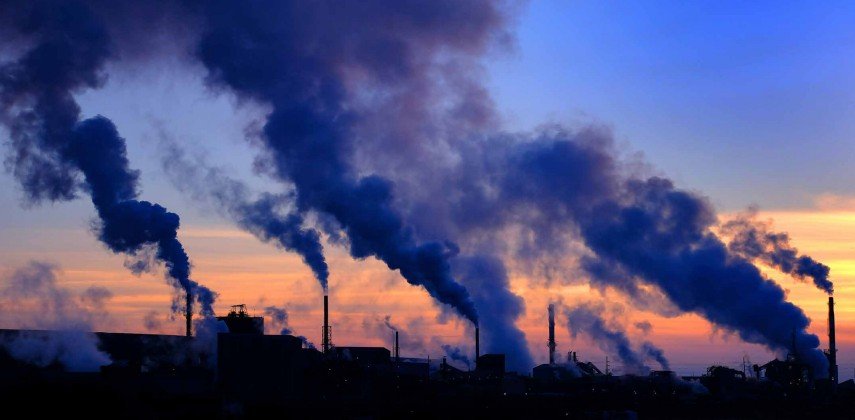 A large factory in silhouette against a clear evening sky with the sunsetting and large plumes of smoke coming out of the factory chimneys.