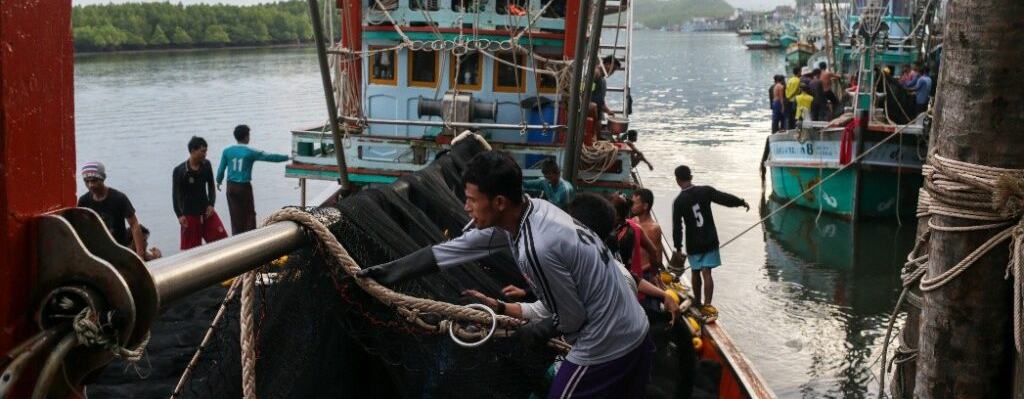 Fishing boats in a port. A man with a cigarette in his mouth is pulling on a thick piece of rope on board one of the boats.