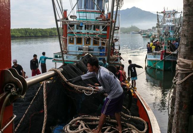 Fishing boats in a port. A man with a cigarette in his mouth is pulling on a thick piece of rope on board one of the boats.