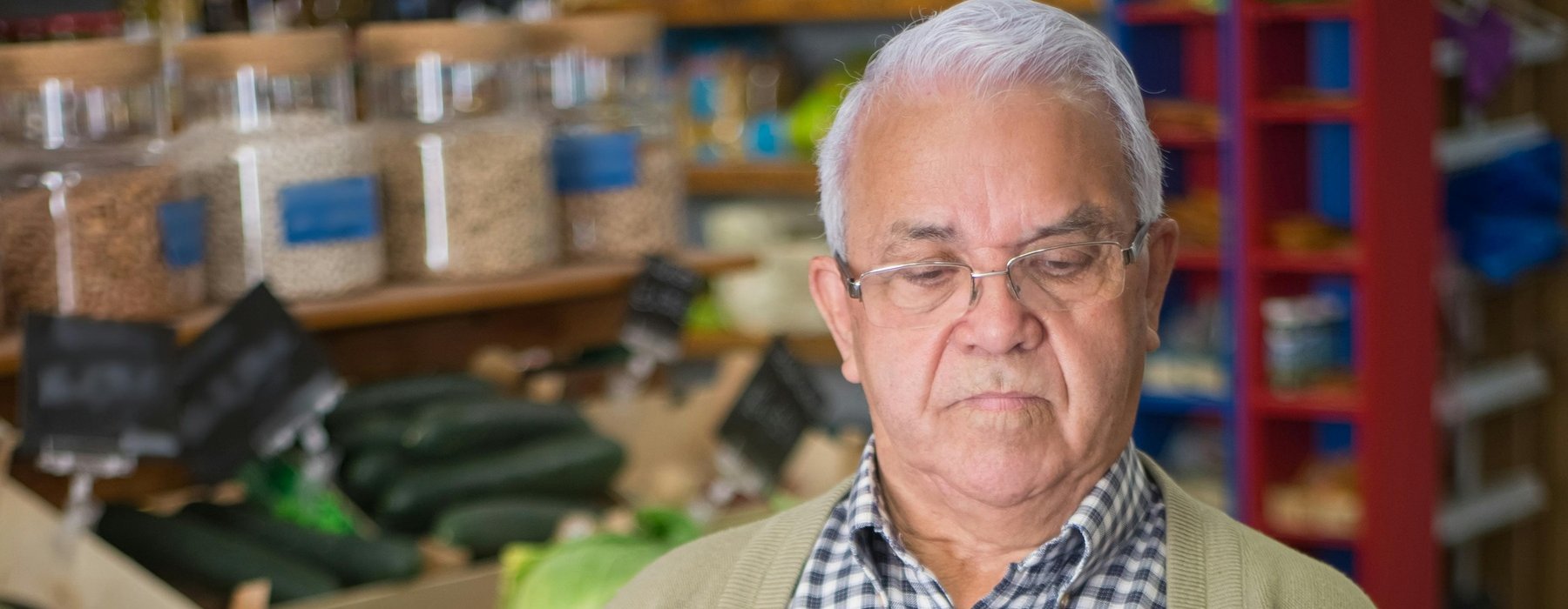 An older man wearing a navy checked shirt and a pale cardigan is holding a basket of produce in a shop whilst looking at the back of a box