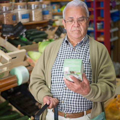 An older man wearing a navy checked shirt and a pale cardigan is holding a basket of produce in a shop whilst looking at the back of a box